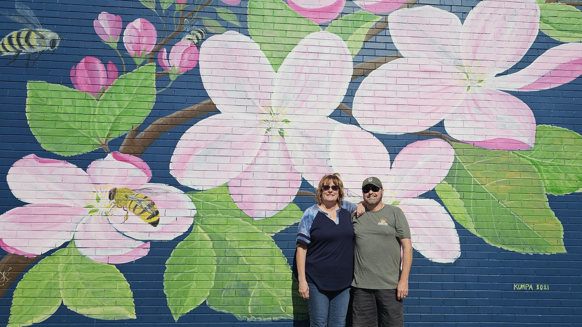 A woman and man smiling in front of a pink, green, and blue floral mural as they participate in the mural challenge