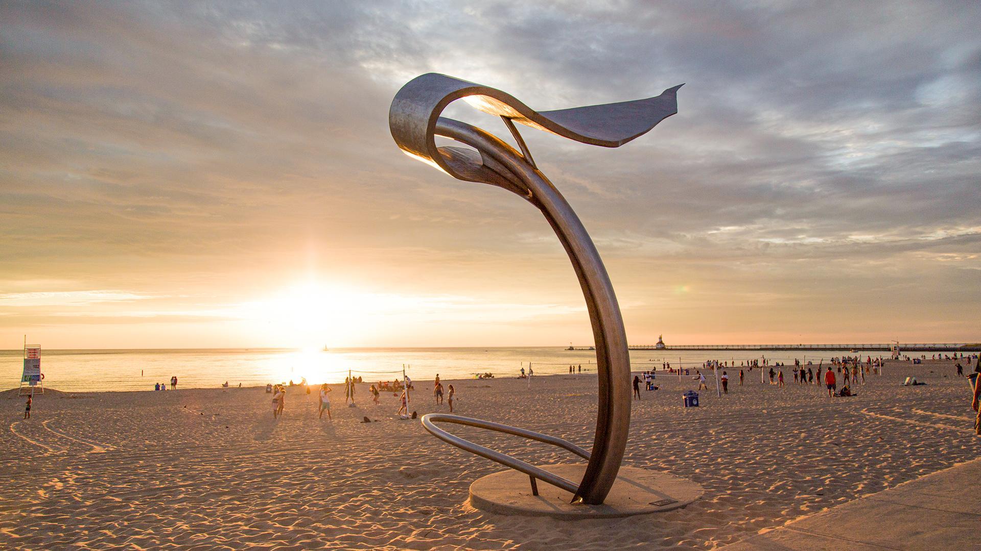 A sculpture called Young Flight by Gail Mally-Mack on Silver Beach, with people enjoying the summer weather and the sunset beginning on the horizon in the background
