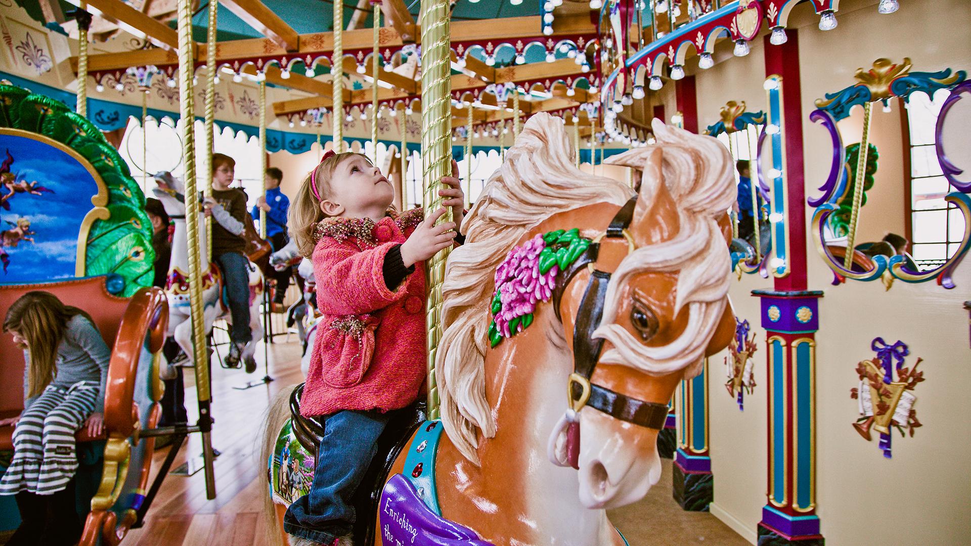 A young girl looking up while riding the Silver Beach Carousel, with other children on carousel horses in the colorful background