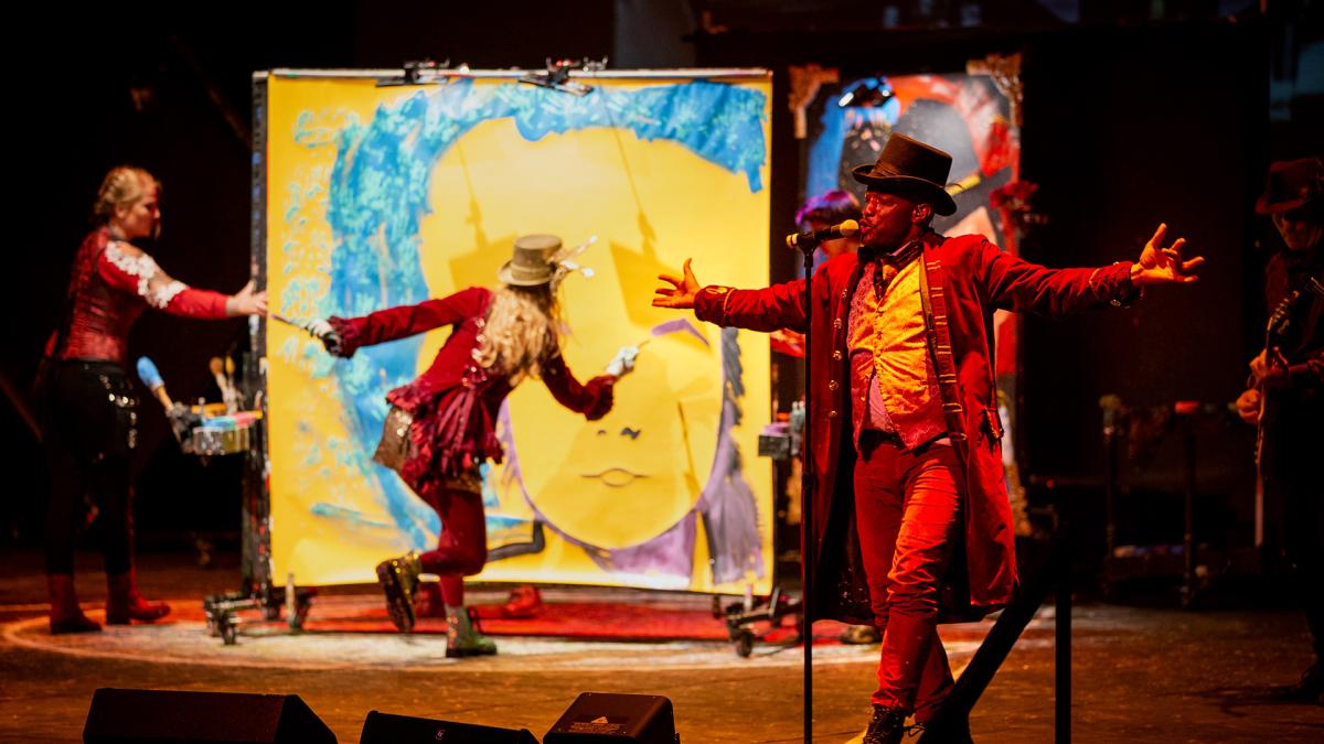On stage at the Mendel Center, a man in a red coat and top hat stands at a microphone with arms outstretched under stage lighting, while two costumed performers in the background paint a face on a large canvas.