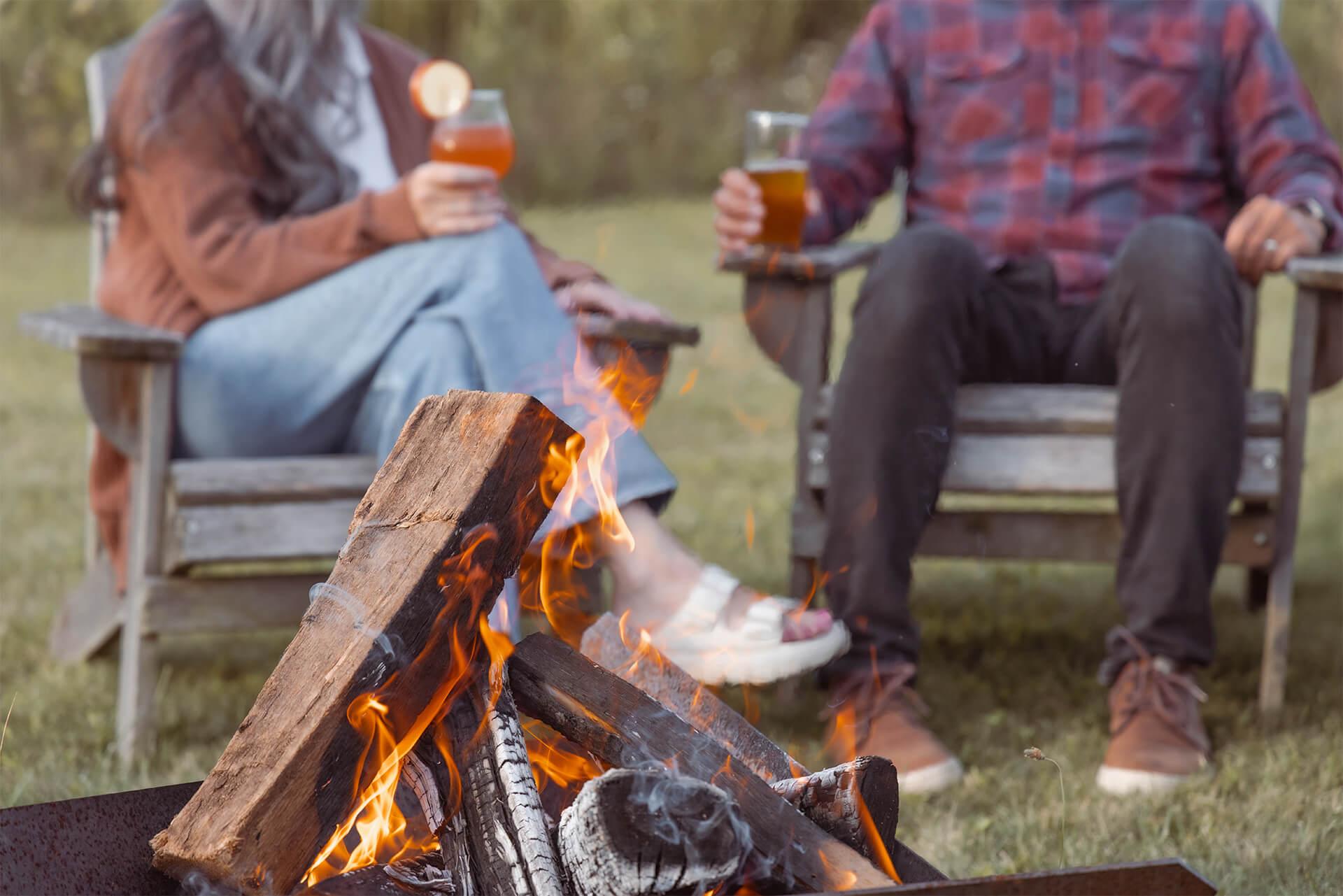 Two people sit in wooden chairs, holding drinks, near a campfire with burning logs. The scene conveys warmth and relaxation in an outdoor setting.