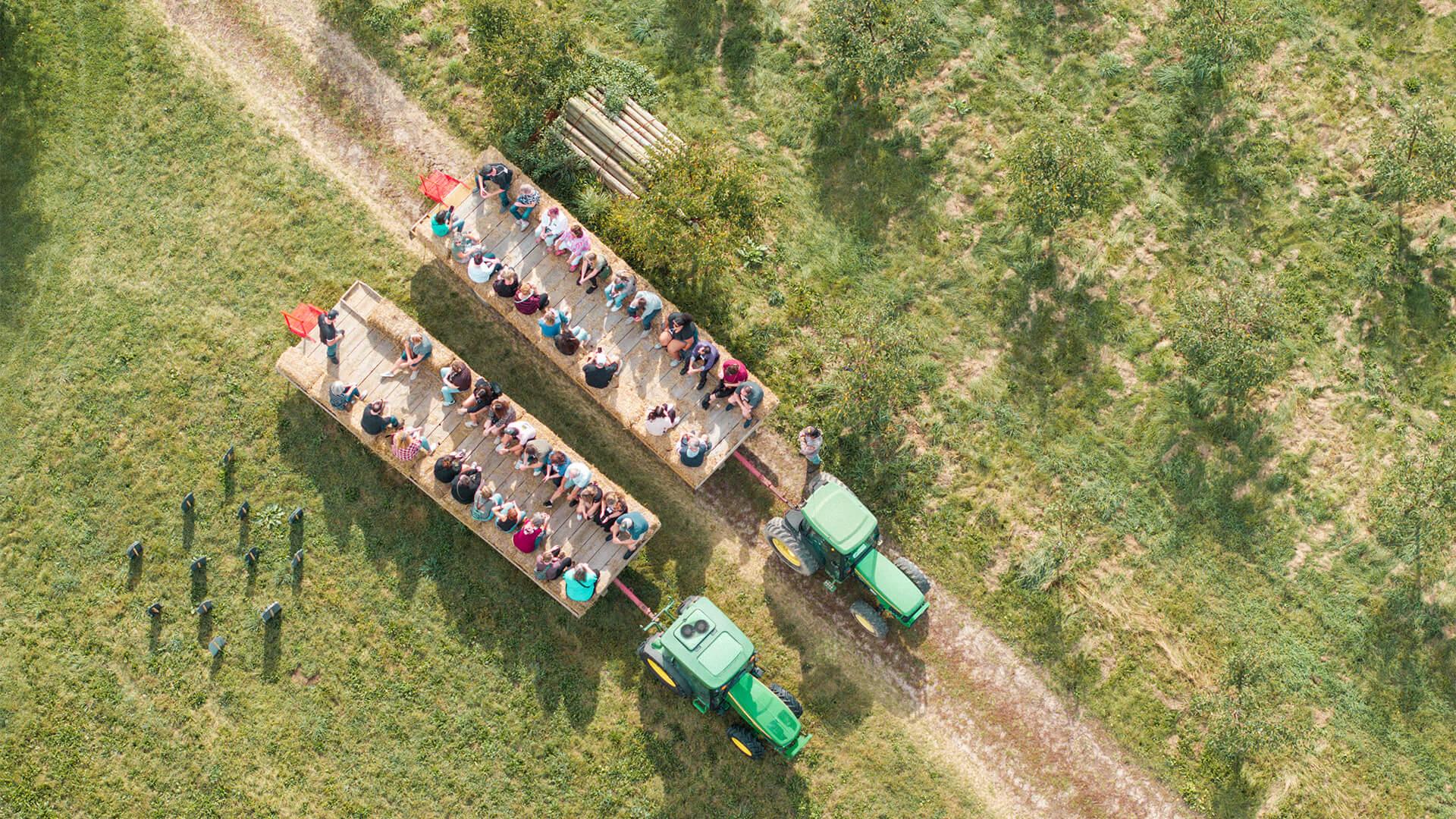 Aerial view of two green tractors pulling wooden trailers with seated people along a path through a lush, grassy orchard, evoking a relaxed, pastoral vibe at Jollay Orchards.