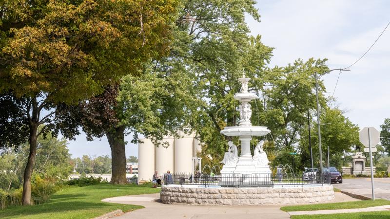 A white, ornate fountain sits in a park surrounded by trees and a stone walkway. The clear sky and greenery create a peaceful and inviting atmosphere.