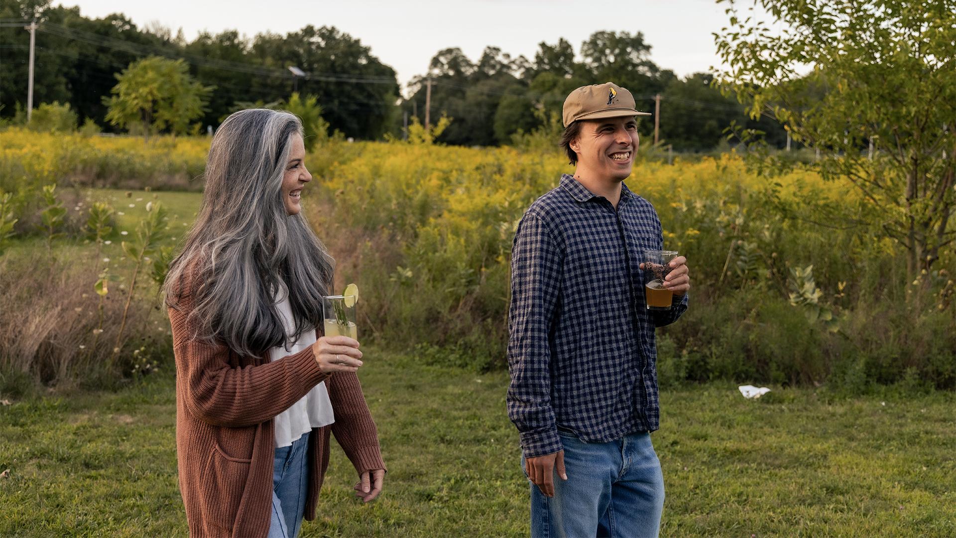 A woman with gray hair and a man in a cap, both smiling and holding drinks, walk through a sunny, grassy field bordered by trees and wildflowers.