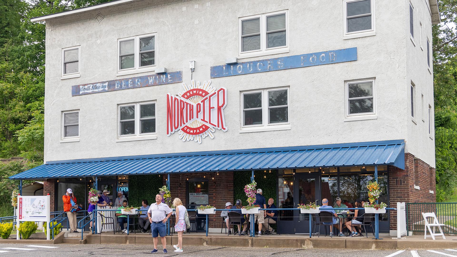 Two-story building with a blue awning and -North Pier- sign. Outdoor seating filled with people dining. Trees in the background add a relaxed vibe.