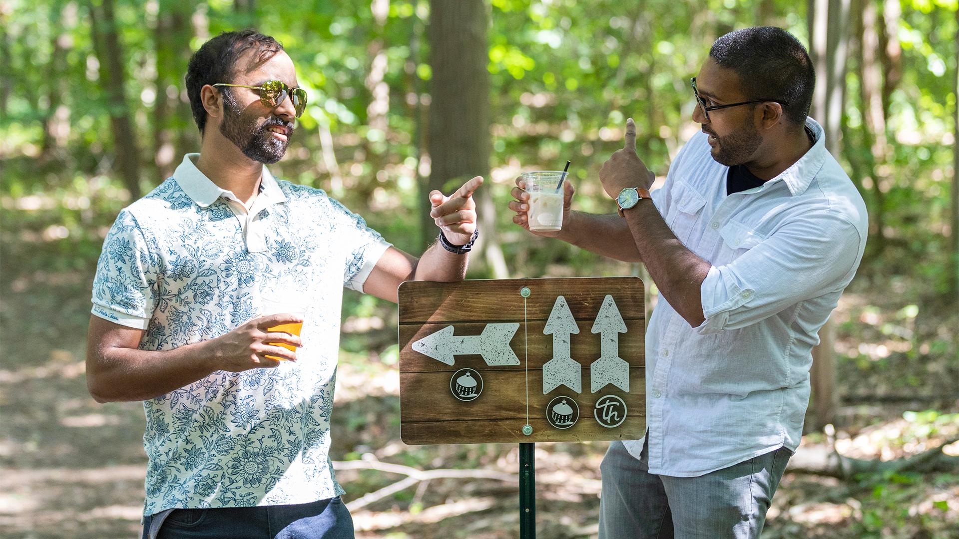 Two men in sunglasses and casual shirts stand outdoors, smiling and pointing at a wooden sign with arrows in a forest setting, conveying a relaxed, joyful mood.