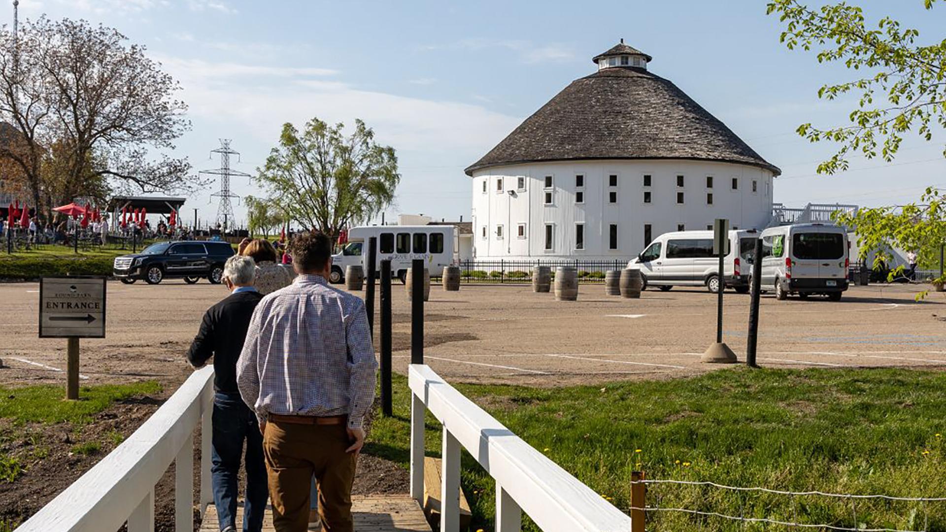 People walk on a path towards a large, white, round barn with a domed roof under a clear sky. The scene is bright and open, with a sense of exploration.