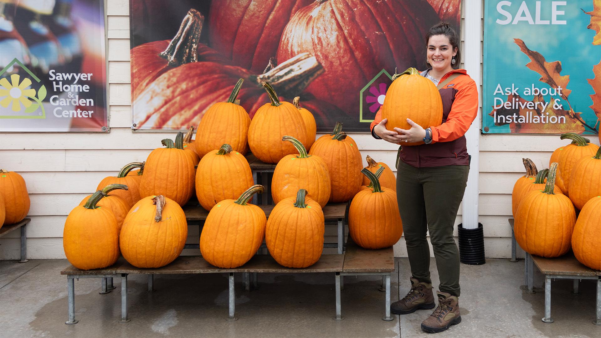 A woman smiling holds a large pumpkin, standing next to a display of pumpkins. Posters are in the background promoting Sawyer Home and Garden Center.