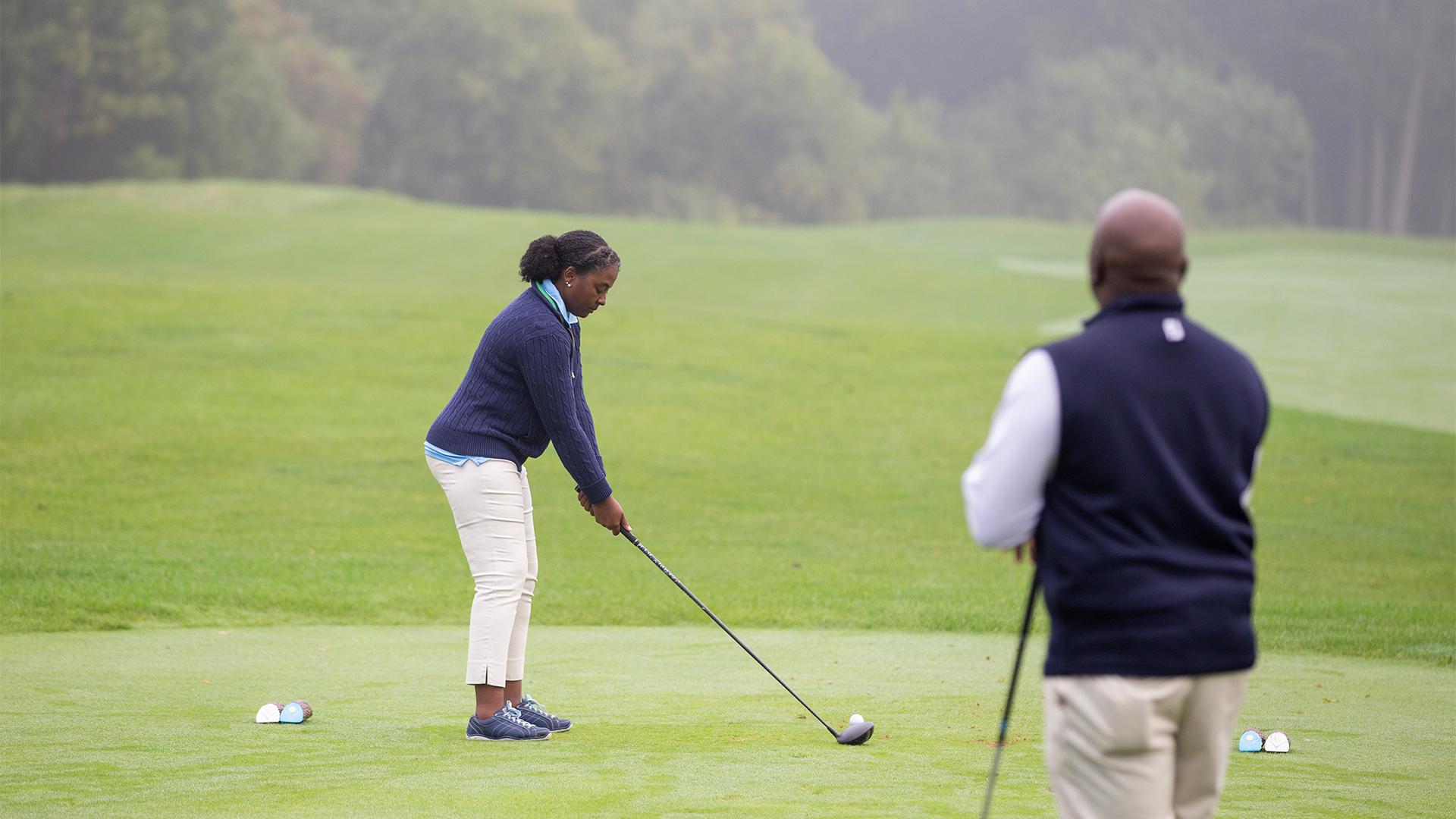 A woman golfer prepares to drive on a lush, green course, holding a club, while another person watches. The setting is calm with misty trees in the background.