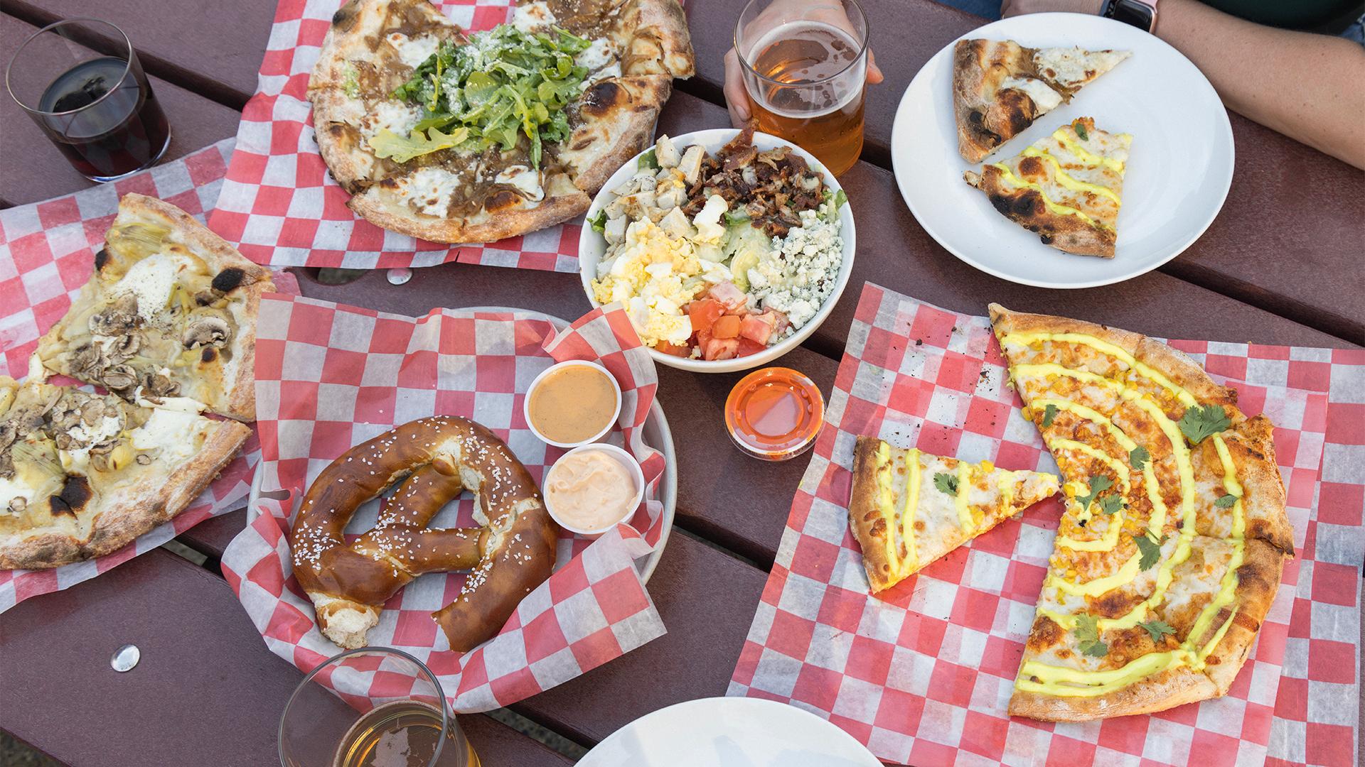A table with various foods on red checkered paper, including pizzas, a pretzel with dips, and a salad bowl at Haymarket. Drinks in glasses suggest a casual, social meal.