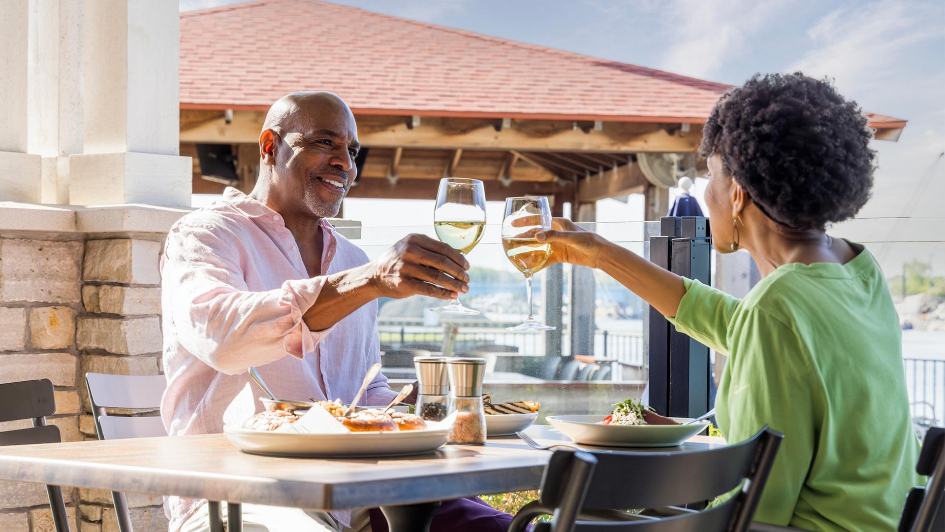 A smiling couple clinks glasses of white wine at an outdoor restaurant, with sunny weather and waterfront views, conveying warmth and enjoyment.