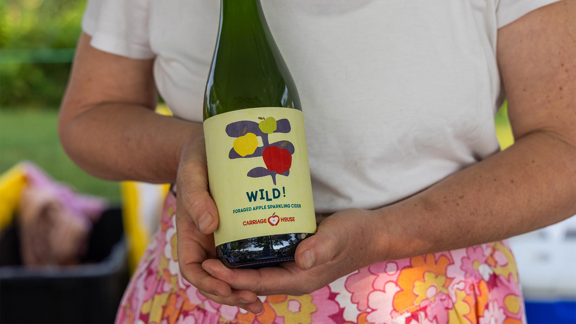 A person in a floral skirt holds a green bottle labeled -Wild! Foraged Apple Sparkling Cider,- with colorful fruit graphics, against an outdoor backdrop.