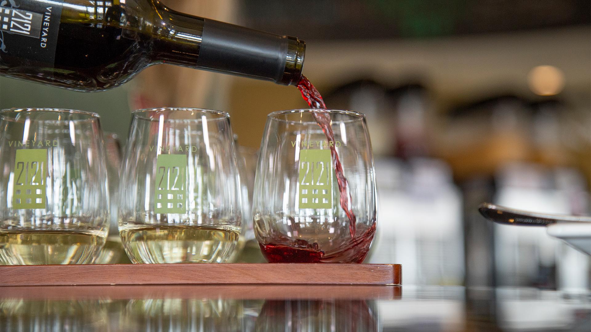 A glass of red wine is being poured beside two glasses of white wine on a wooden tray. The setting is a sophisticated wine tasting bar.