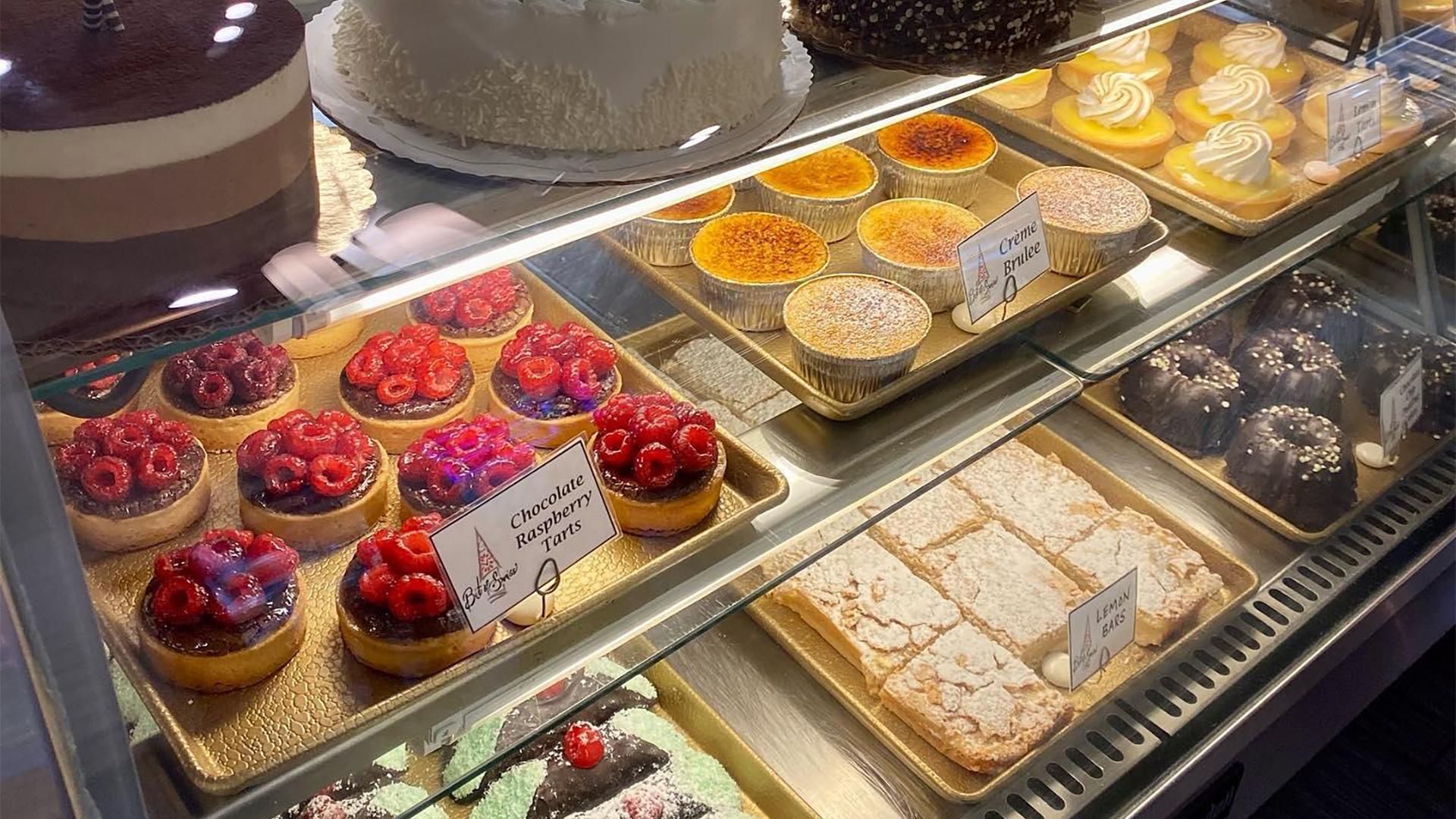 A bakery display case at Bit of Swiss filled with various desserts, including chocolate raspberry tarts, crème brûlée, lemon tarts, square pastries, and dome cakes.