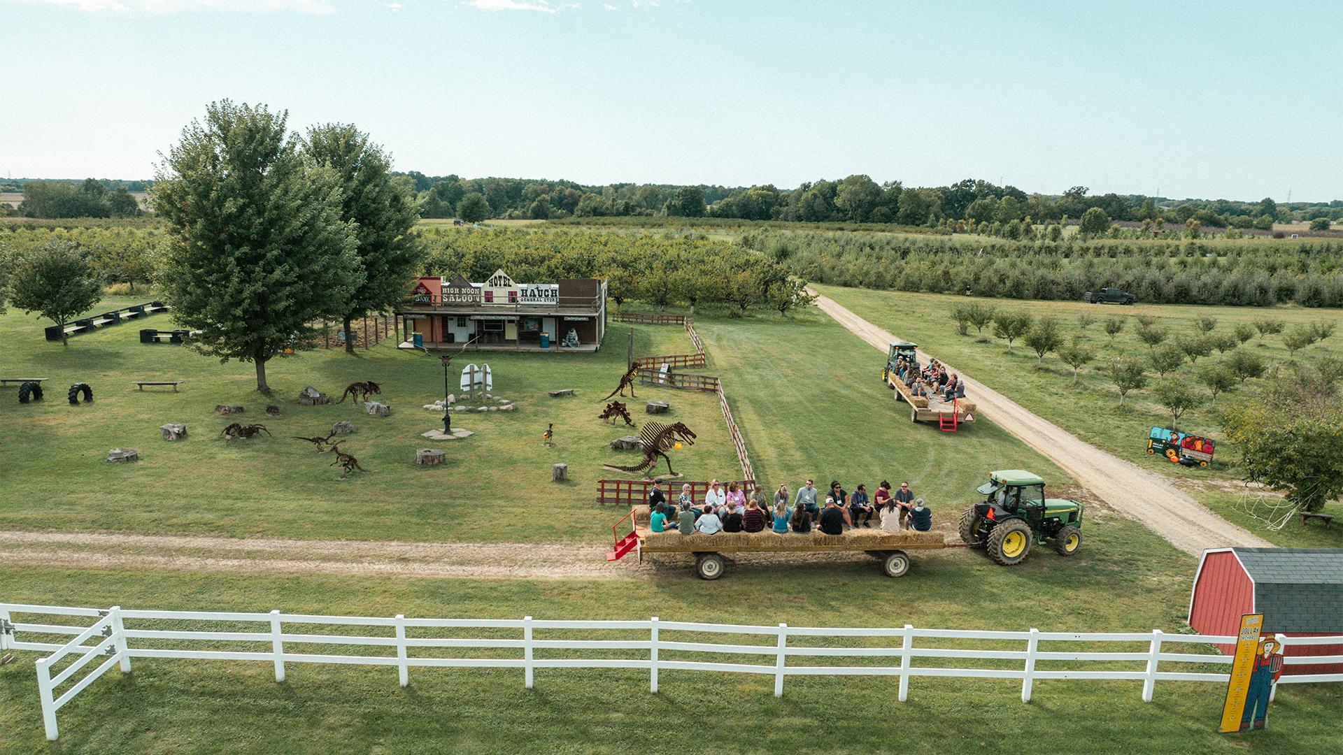 Aerial view of Jollay Orchards with a tractor pulling a hayride wagon filled with people. Nearby are trees, a white fence, and a rustic building