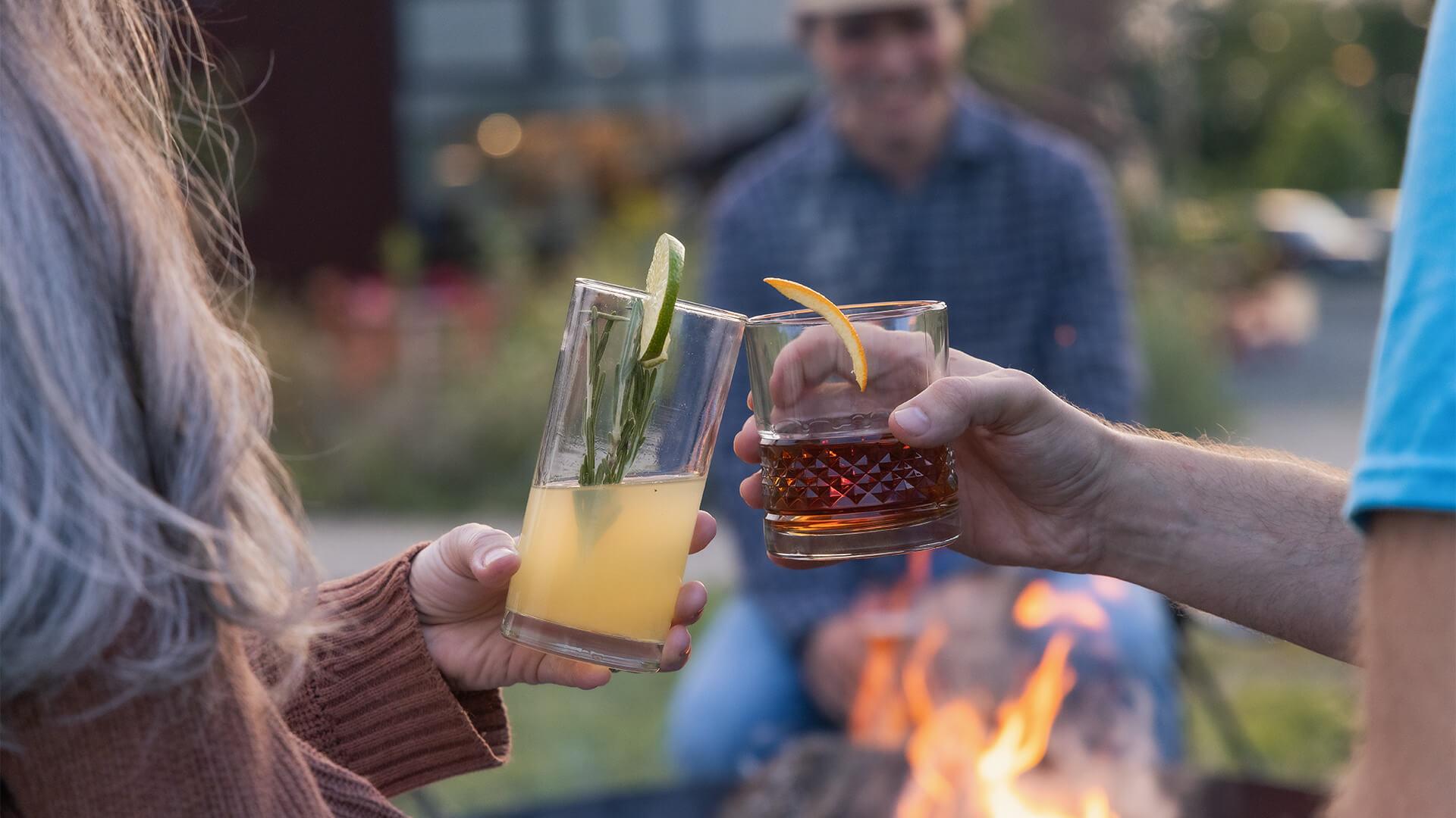 Two people toast with cocktails near a campfire. One holds a clear glass with a yellow drink and lime, the other a glass with dark liquid and an orange peel. In the background, a person sits, blurred. The scene conveys a warm and relaxed mood.