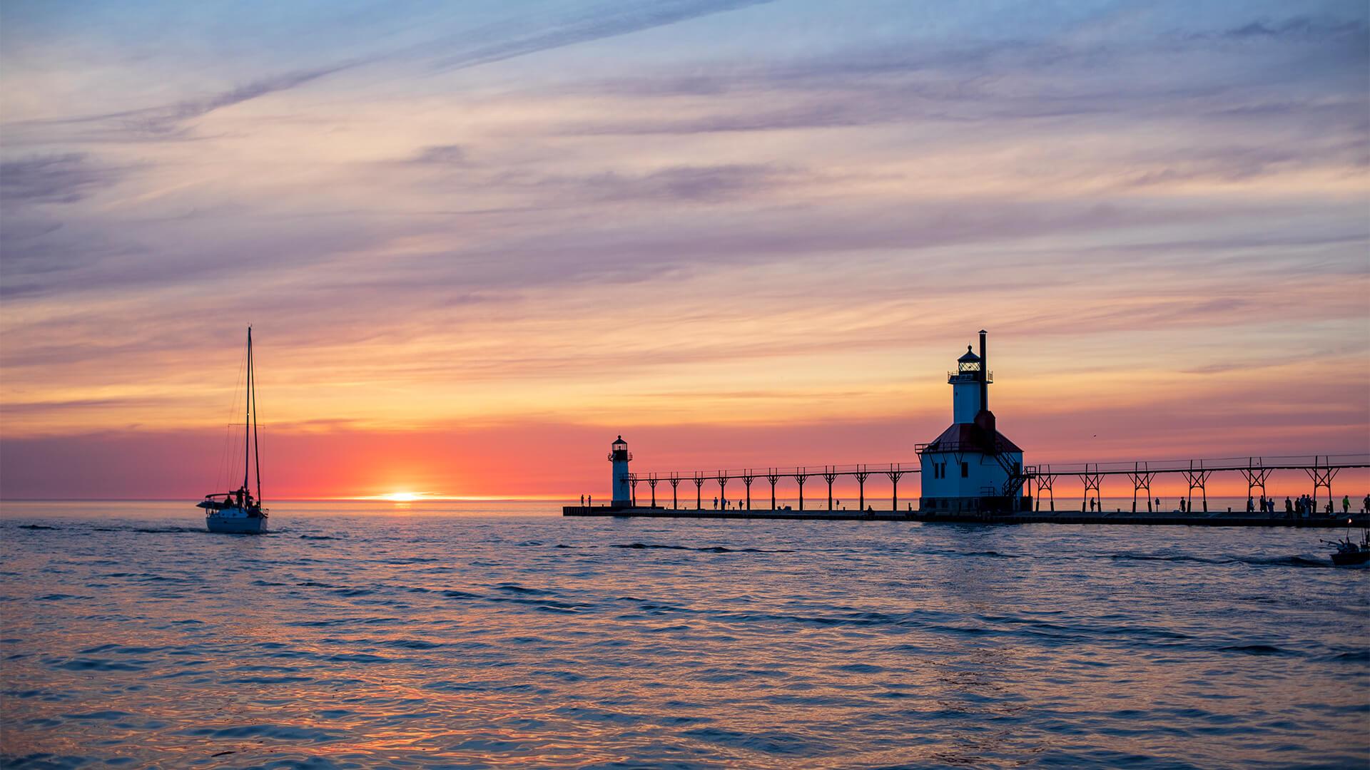 Serene sunset over a calm sea with a lighthouse and pier silhouetted, a sailboat on the left. The sky glows orange and purple, evoking tranquility.