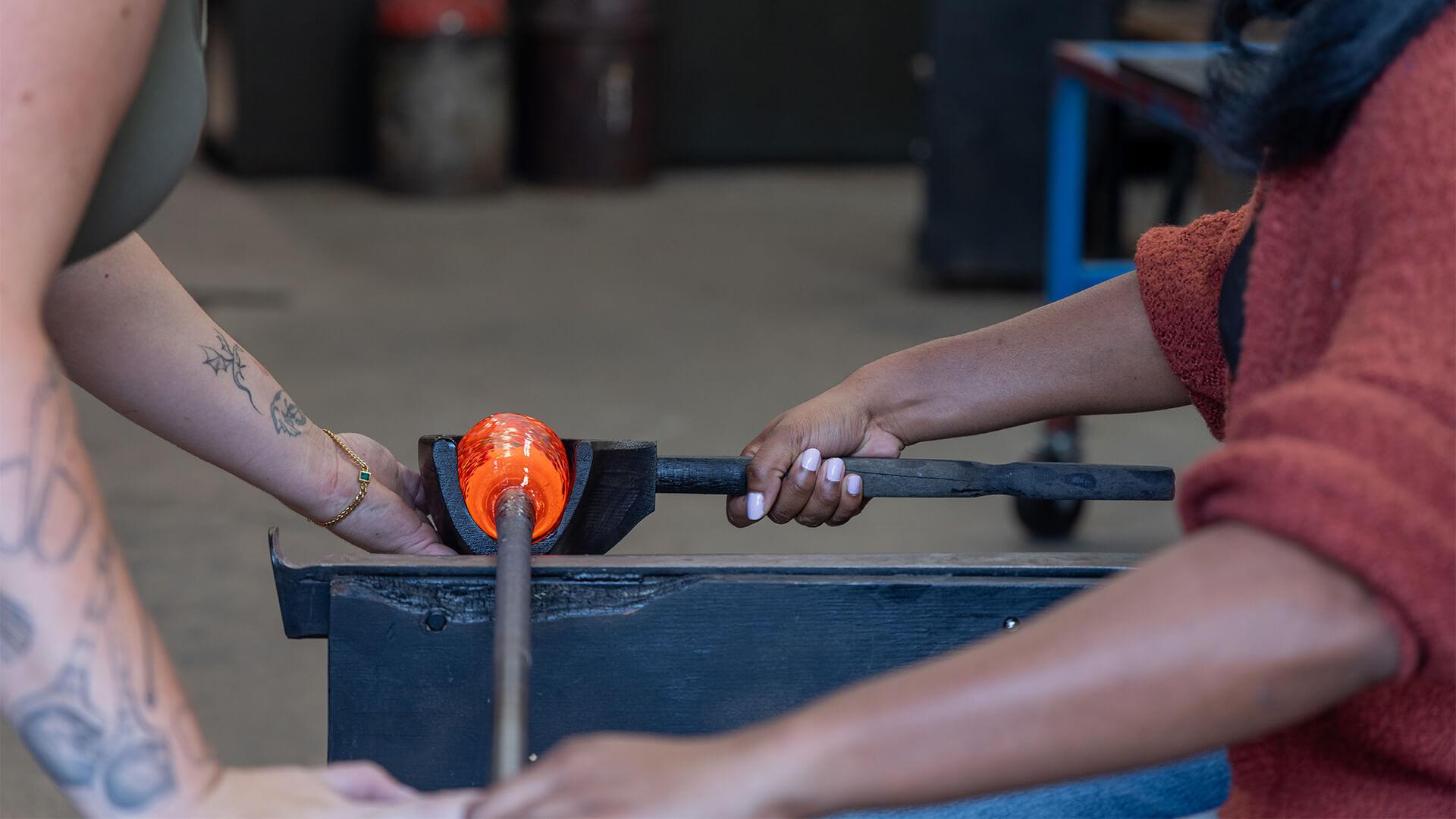 Two people collaborate on glassblowing. One holds a rod with glowing hot glass, while the other assists. The workshop setting conveys focus and craftsmanship.
