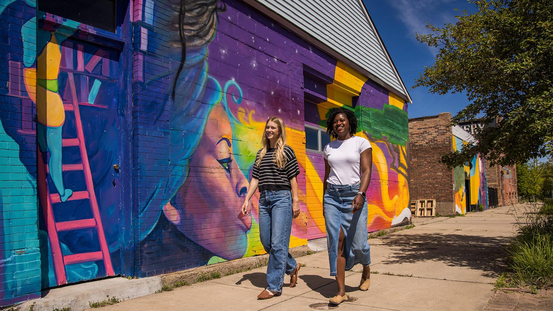 Two women walk along a sidewalk beside a colorful mural featuring a large woman