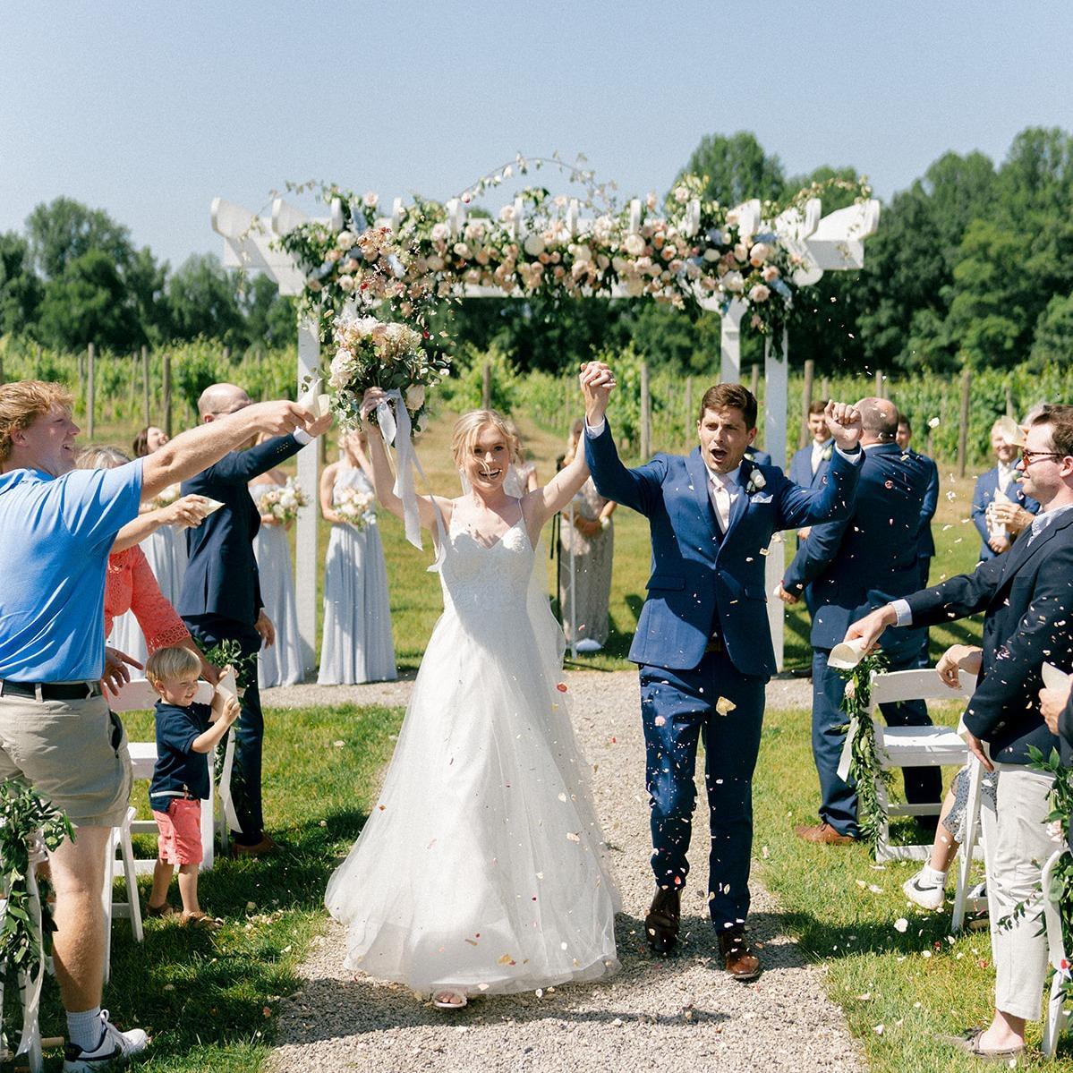A joyful bride and groom in wedding attire walk hand in hand under a floral arch outdoors, surrounded by guests throwing confetti on a sunny day.
