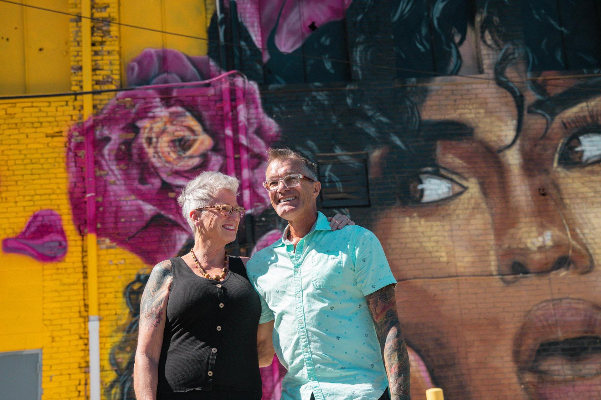 Two people posing infront of the Cornerstone Alliance Building mural. 