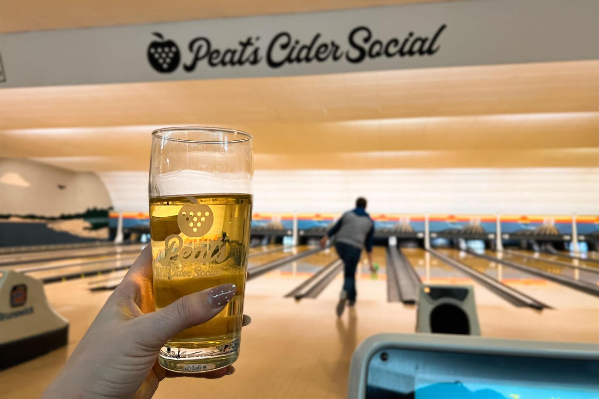 A person holds up a glass of cider with bowling action in the background.