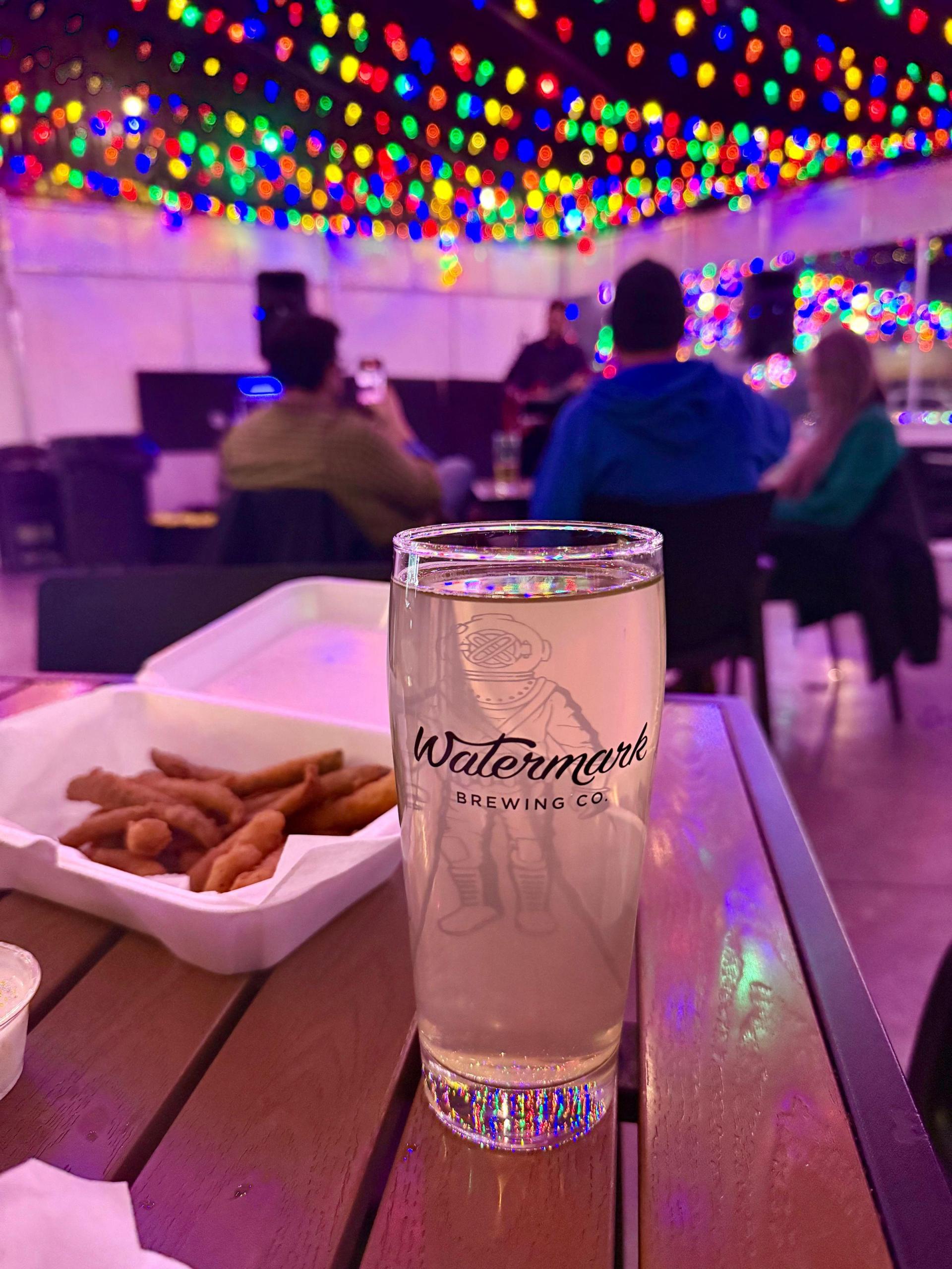 A pint of beer inside Watermark Brewing Company, with the interior decorated with holiday lights.