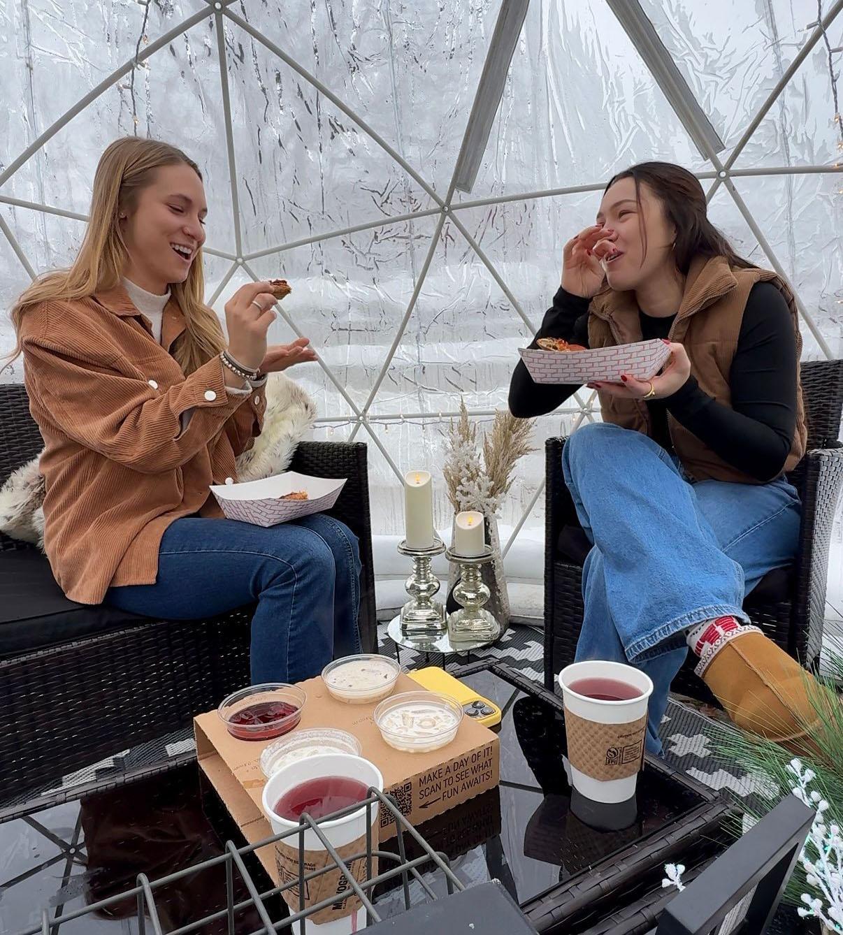 Two people relax with drinks and food inside a cozy igloo at Round Barn Estate.