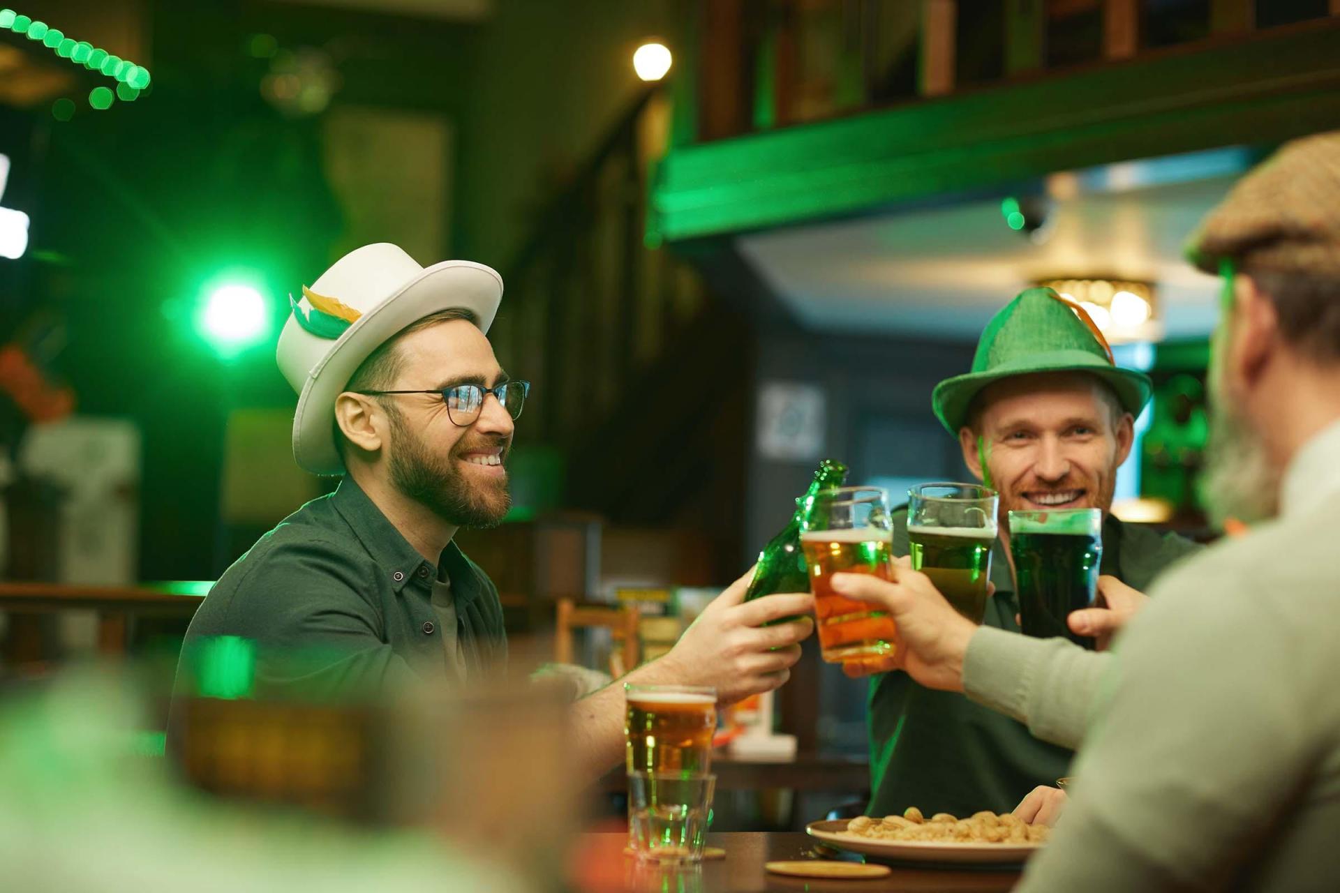 Three friends in festive hats cheer with green and amber drinks in a dimly lit pub, smiling warmly, creating a joyful and celebratory atmosphere.
