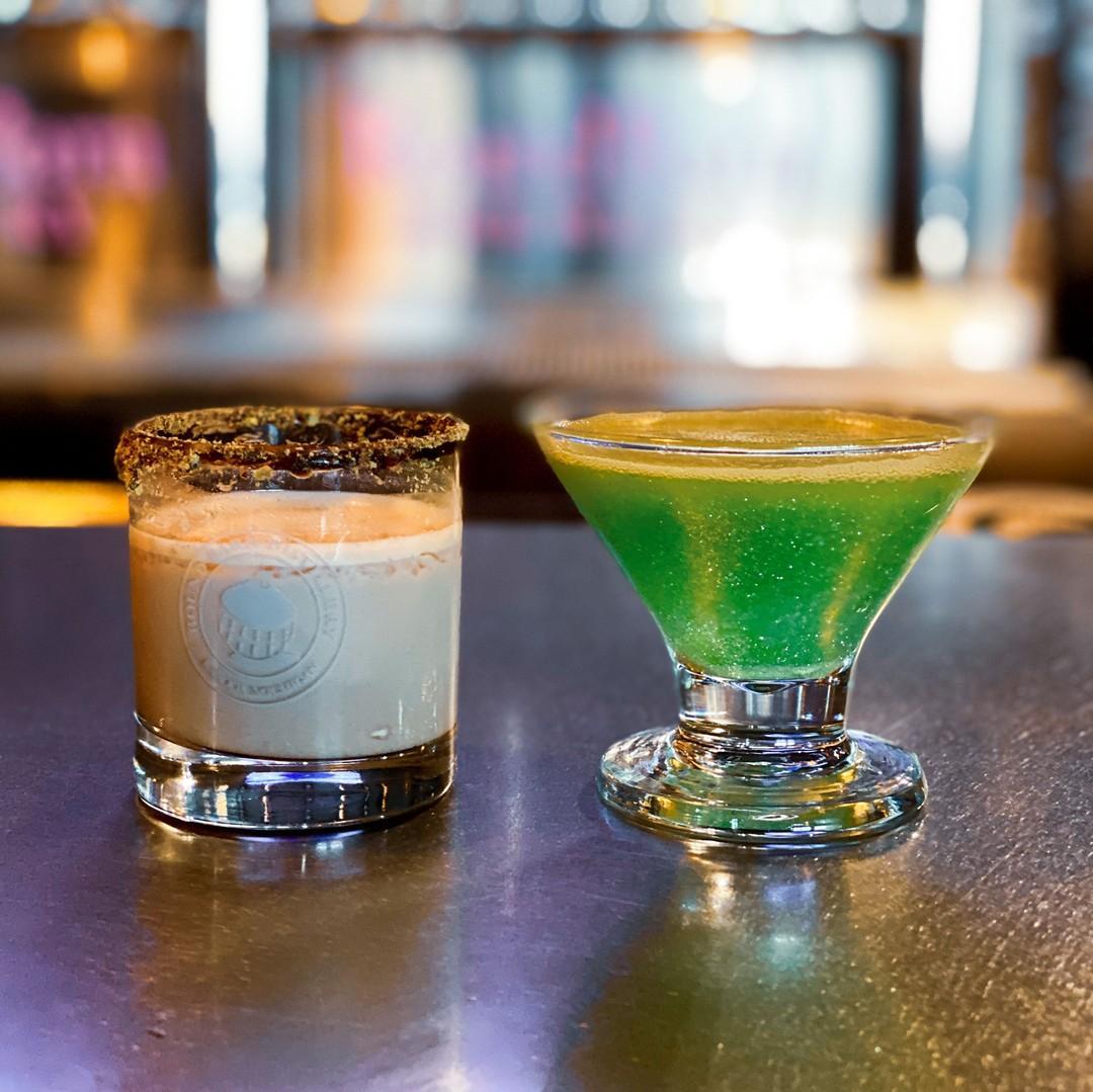 A creamy beige cocktail with a chocolate-rimmed glass sits beside a vibrant green, frosty drink on a reflective bar counter at Round Barn Brewery & Public House.