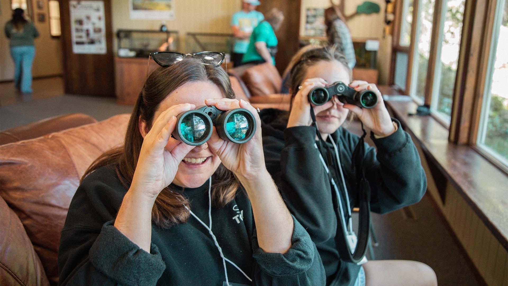 Two people using binoculars to watch birds from the indoor bird viewing room at Sarett Nature Center.