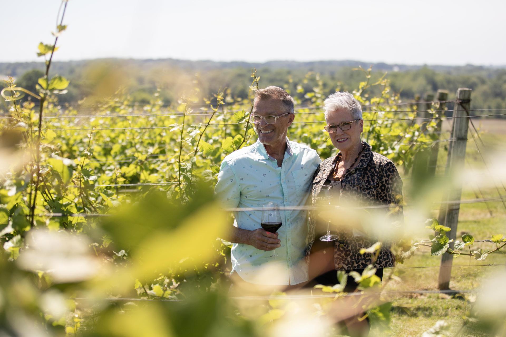 A couple standing in a vineyard holding glasses of wine.
