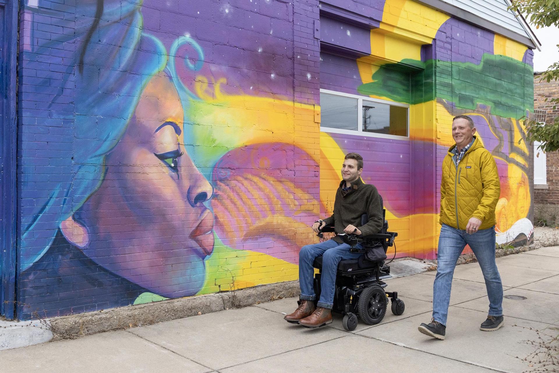 Two people explore the Benton Harbor Arts District, one traveling in a wheelchair, as they pass a vibrant mural.